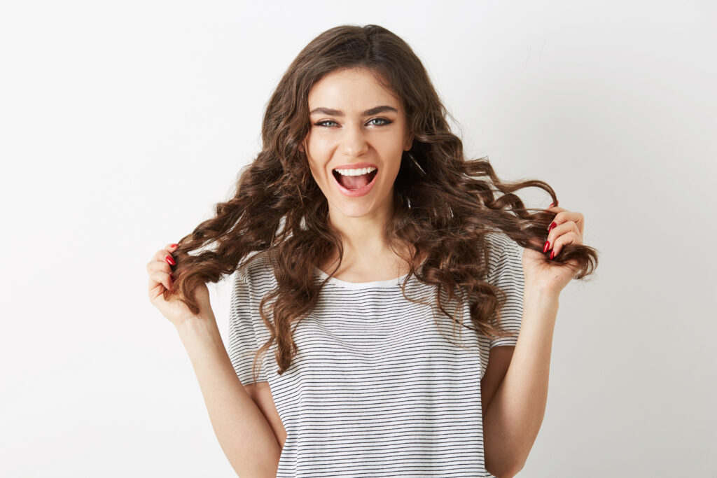 attractive young lady holding her long brown curly hair laughing with positive face expression, emotional woman, looking in camera, isolated, happy emotion