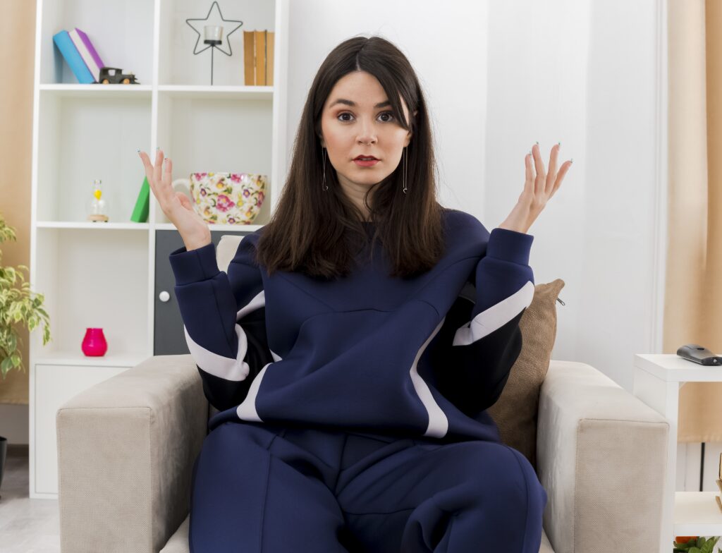 clueless young pretty caucasian woman sitting armchair designed living room looking keeping hands air