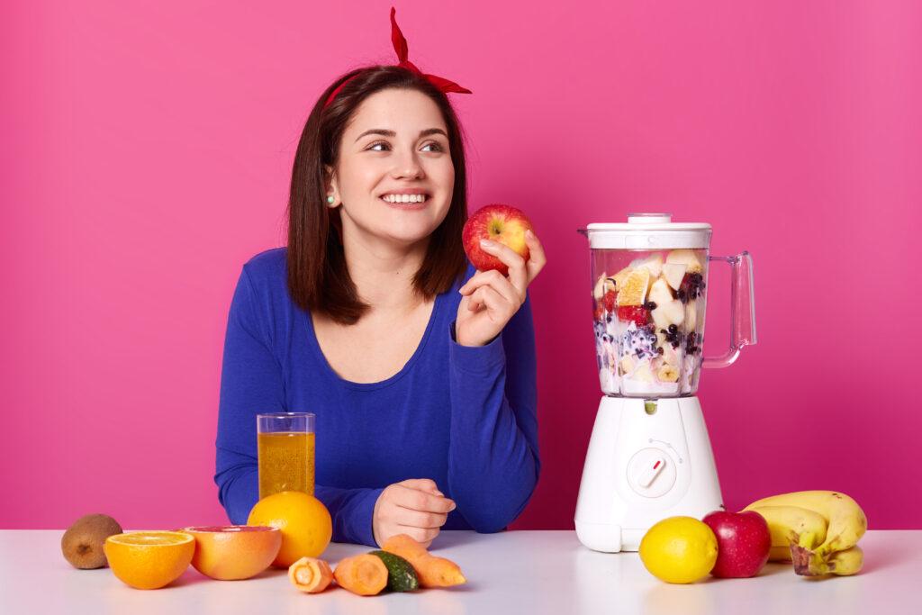 smiling girl with fresh fruits on table isolated over pink background. lady looks aside while making coktail. woman holds apple in left hand thinks about something pleasent. health concept.