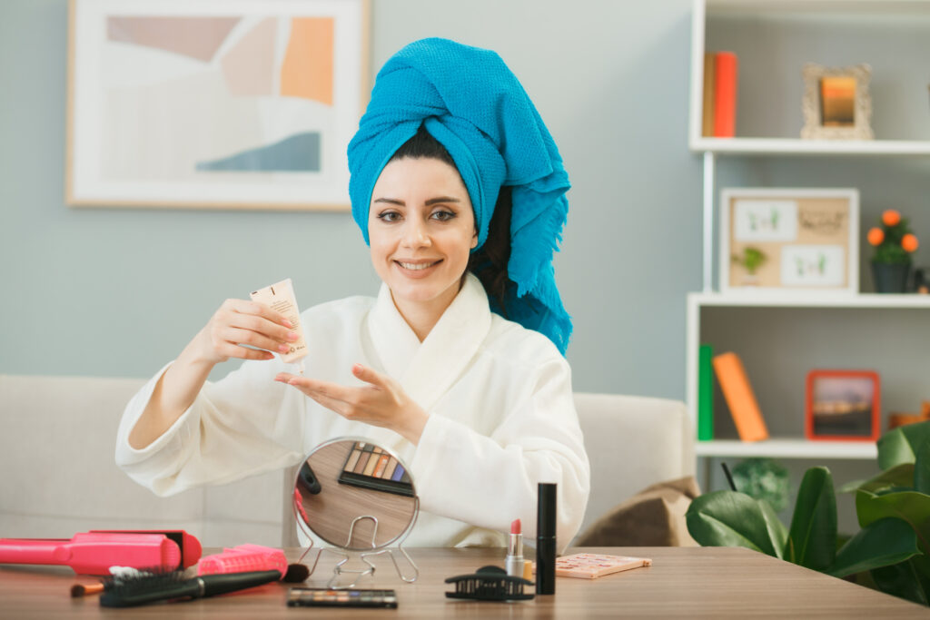 smiling looking camera young girl holding tone up cream wrapped hair in towel sitting at table with makeup tools in living room