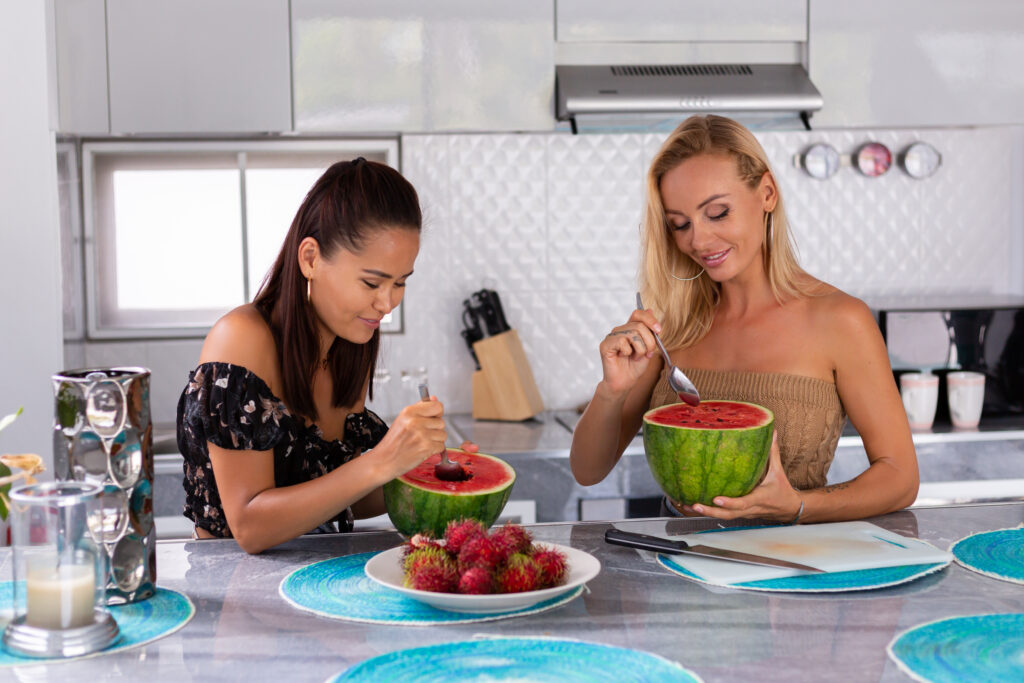 two girl friends eating watermelon rambutan tropical fruits kitchen